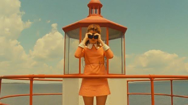 Fotograma de Moonrise Kingdom, de Wes Anderson: menina de vestido laranja com binóculos num farol, paleta pastel